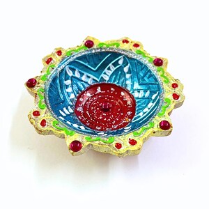 May include: A decorative diya oil lamp featuring a scalloped edge painted in yellow with green accents and small red beads. The interior bowl is painted in bright blue with white floral patterns, while the center is a solid red circular base.
