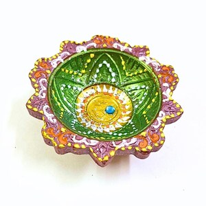 May include: A hand-painted clay diya lamp featuring a scalloped edge with purple, orange, and white details. The interior bowl is painted green with white floral patterns and a central yellow circle containing a blue bead and an Om symbol.