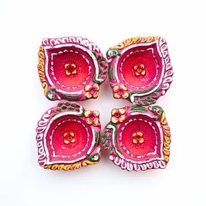 May include: Four hand-painted clay diya lamps arranged in a square formation. Each lamp features a vibrant pink interior with a yellow floral center, surrounded by decorative white, green, and yellow painted patterns along the scalloped edges.