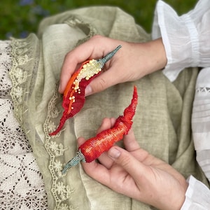 May include: Two red and orange beaded pepper-shaped ornaments with green and gold accents. The ornaments are held in the hands of a person.