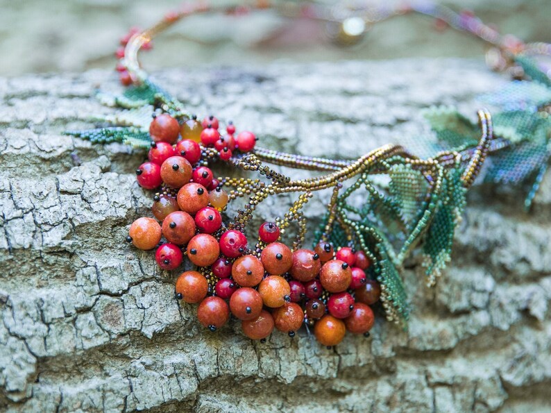 May include: A beaded necklace with a leafy design and red berries. The necklace is made with small beads and is a beautiful example of handmade jewelry.