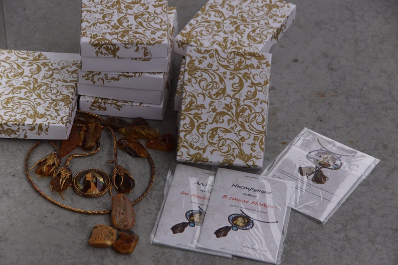 May include: A collection of amber necklaces and earrings displayed on a table with white boxes and instructions in Russian. The jewelry is packaged in white boxes with a gold floral pattern. The instructions are printed on white paper with a clear plastic cover.