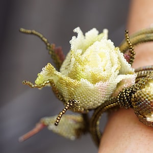 Może przedstawiać: Ręcznie robiona bransoletka z różą wykonana z koralików. Róża jest w kolorze złamanej bieli z żółtymi i różowymi odcieniami, łodyga i liście są zielone i brązowe. Szczegółowy, kwiatowy dodatek.