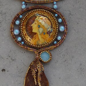 May include: A beaded necklace with a large pendant featuring a painted portrait of a woman with a crown and a blue stone. The pendant is surrounded by blue beads and the necklace is made of brown and gold beads. The pendant is attached to a long amber-colored stone.