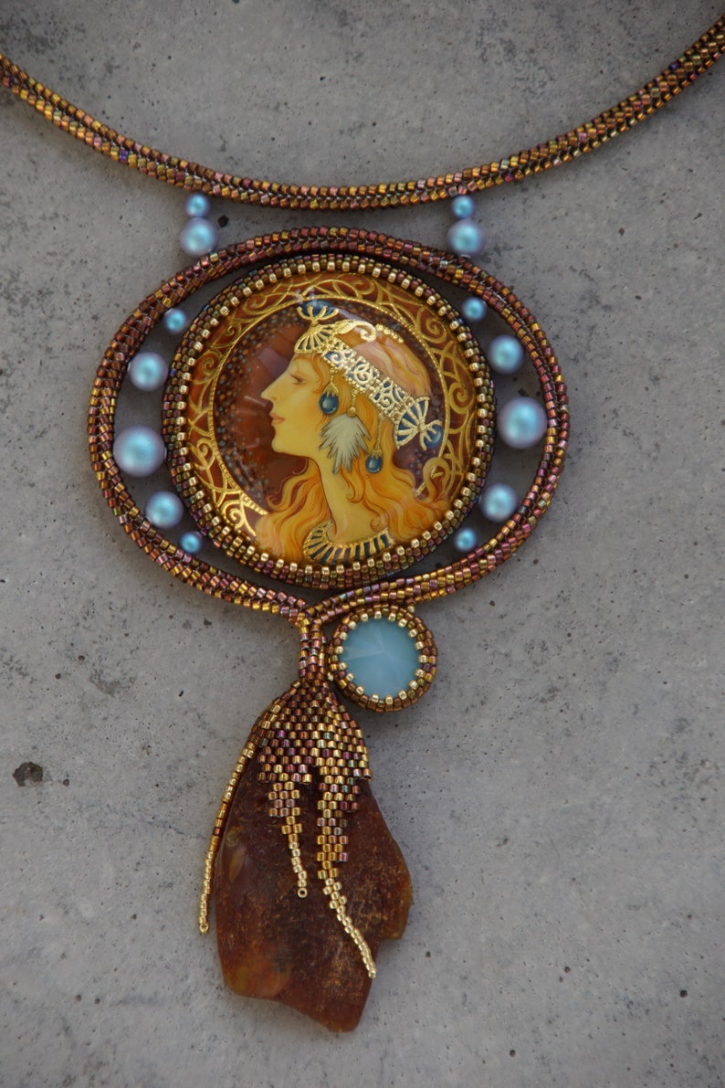 May include: A beaded necklace with a large pendant featuring a painted portrait of a woman with a crown and a blue stone. The pendant is surrounded by blue beads and the necklace is made of brown and gold beads. The pendant is attached to a long amber-colored stone.