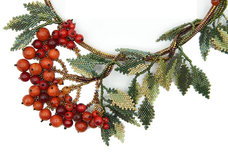 May include: A beaded necklace with a cluster of red and orange berries and green leaves. The necklace is made of small beads and is a beautiful example of beadwork.