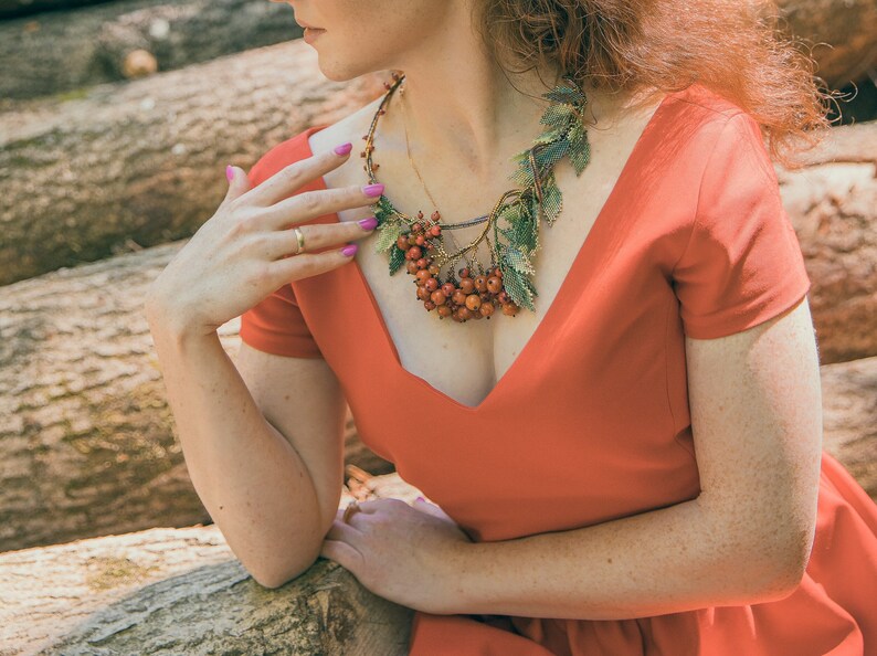 May include: A woman wearing a red dress with a unique necklace made of leaves and red berries.