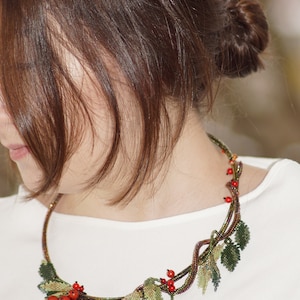 May include: A beaded necklace with a design of a branch with red berries and green leaves. The necklace is worn by a person with brown hair.