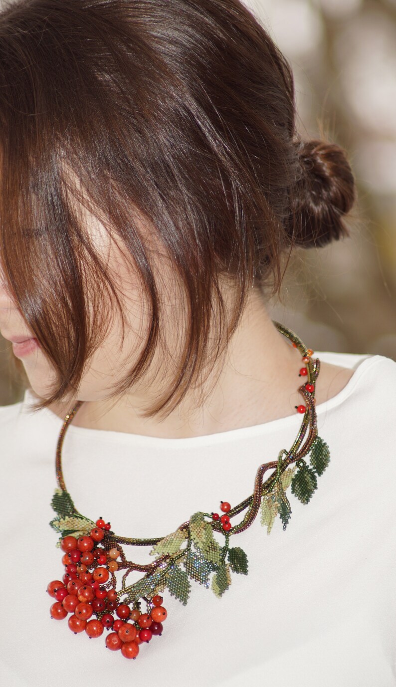 May include: A beaded necklace with a design of a branch with red berries and green leaves. The necklace is worn by a person with brown hair.
