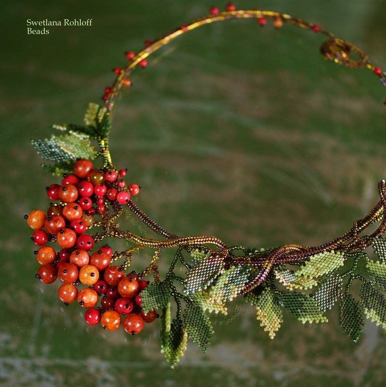 May include: A beaded necklace with a design of a branch with red berries and green leaves. The necklace is made of small beads and is a beautiful piece of jewelry.