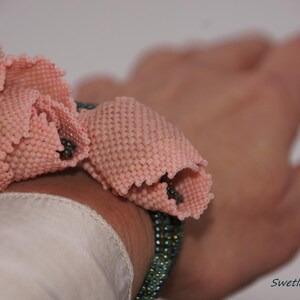 Könnte beinhalten: Ein rosa Perlenarmband mit Blumenmuster. Das Armband besteht aus kleinen Perlen und wird am Handgelenk getragen.