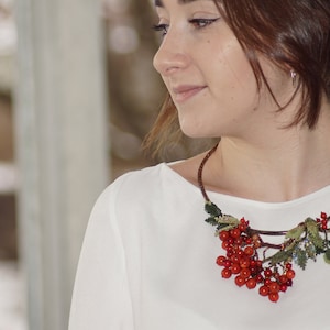 May include: A woman wearing a white shirt with a red berry and green leaf necklace. The necklace is made of beads and wire and is shaped like a branch.