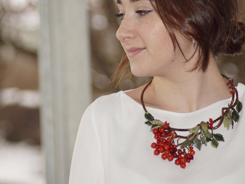 May include: A woman wearing a white shirt with a red berry and green leaf necklace. The necklace is made of beads and wire and is shaped like a branch.