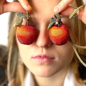 以下が含まれることがあります： 女性がビーズでできたリンゴのイヤリングを着用しています。イヤリングは赤とオレンジで、緑の葉が付いています。