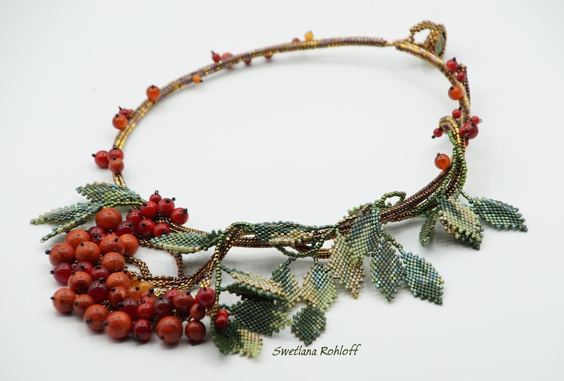 May include: A beaded necklace with a design of leaves and red berries. The necklace is made with brown, green, and red beads. The necklace is on a white background. "Svetlana Rohloff" is written in the bottom right corner of the image.