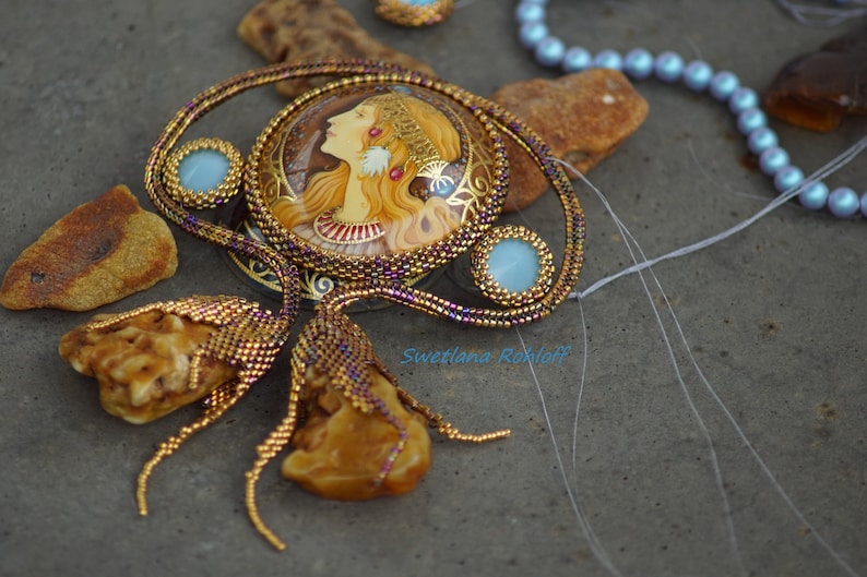 May include: A gold and purple beaded necklace with a round pendant featuring a painted portrait of a woman with long blonde hair. The pendant is surrounded by smaller beads and a light blue stone. The necklace is on a gray surface with amber stones.