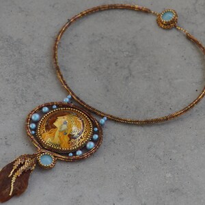 May include: A gold beaded necklace with a large pendant featuring a woman's portrait in a gold frame. The pendant is surrounded by small blue beads. The necklace has a brown and gold color scheme.