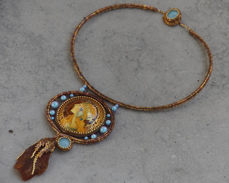 May include: A gold beaded necklace with a large pendant featuring a woman's portrait in a gold frame. The pendant is surrounded by small blue beads. The necklace has a brown and gold color scheme.