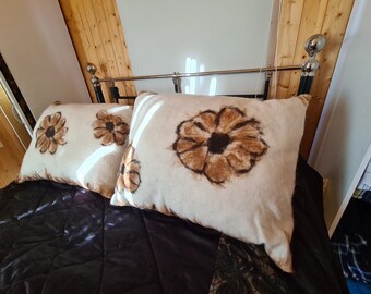 Op de afbeelding: Twee vierkante kussens met een crèmekleurige achtergrond en grote bruine bloemmotieven. De kussens liggen op een donkerpaars sprei. Het bedframe is zilver en zwart.