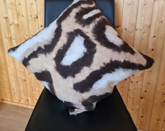 Op de afbeelding: Een vierkant decoratief kussen met een bruin, beige en wit dierenprintpatroon. Het kussen ligt op een zwarte stoel, tegen een houten achtergrond.