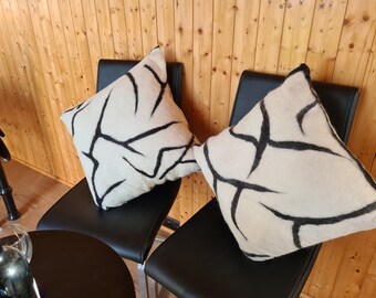 Op de afbeelding: Twee vierkante kussens met een crèmekleurige achtergrond en een gedurfd zwart abstract ontwerp. De kussens zijn geplaatst op zwarte stoelen, wat wijst op een modern interieurthema.