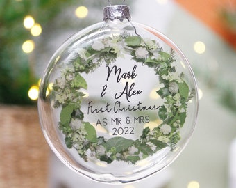 Op de afbeelding: Duidelijke glazen kerstbal met een groene bloemenkrans en de tekst "Mark & Alex First Christmas as Mr & Mrs 2022".