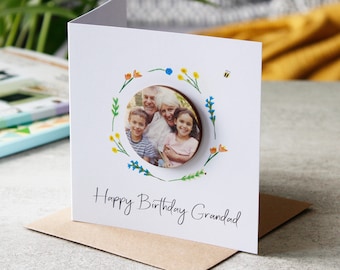 May include: A white greeting card with a family photo in a floral wreath. The card reads "Happy Birthday Grandad" in black script. A small bee illustration is visible. The card is on a brown envelope.