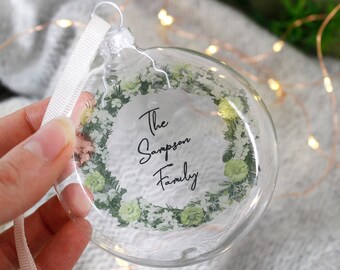 May include: Clear glass Christmas ornament with a white floral wreath design and the text "The Sampson Family" printed in black.