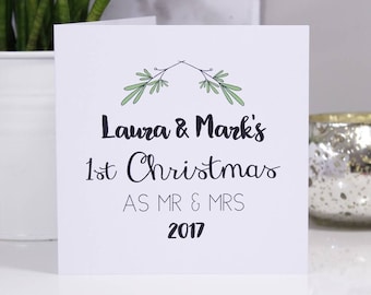 May include: A white greeting card with the text "Laura & Mark's 1st Christmas AS MR & MRS 2017" in black script. The card is decorated with green sprigs at the top. The card is on a white surface.