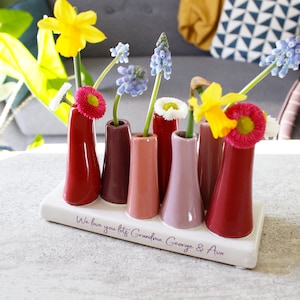 May include: White ceramic vase with six small red, pink and purple bud vases. The vase has the inscription "We love you lots Grandma George & Ava".