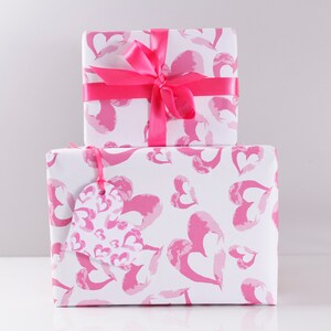 May include: Two gift boxes wrapped in white paper with a pink watercolor heart pattern. The top box has a pink satin ribbon tied in a bow. The bottom box has a pink heart-shaped gift tag.