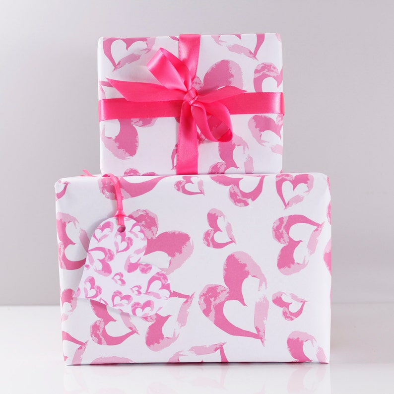 May include: Two gift boxes wrapped in white paper with a pink watercolor heart pattern. The top box has a pink satin ribbon tied in a bow. The bottom box has a pink heart-shaped gift tag.