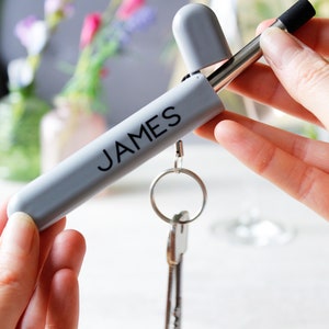 Puede incluir: Una pajita de metal gris con una punta negra, en una funda gris con el nombre "JAMES" impreso en ella. La funda tiene un llavero.