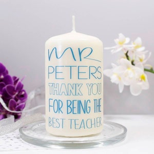 Op de afbeelding: Witte kaars in de vorm van een pilaar met blauwe tekst die "Mr. Peters Bedankt dat je de beste leraar bent" zegt.