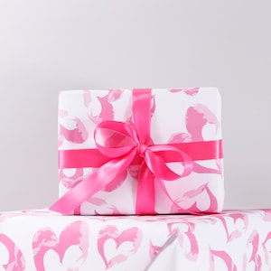 May include: A white gift box with a pink and white heart pattern wrapped with a pink satin ribbon. The box is sitting on a pink and white heart patterned wrapping paper.