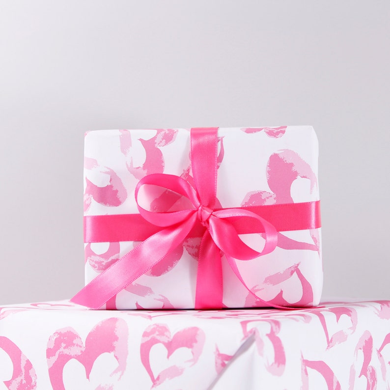 May include: A white gift box with a pink and white heart pattern wrapped with a pink satin ribbon. The box is sitting on a pink and white heart patterned wrapping paper.