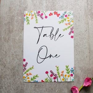 May include: White card with a floral border in shades of pink, yellow, and blue. The card says "Table One" in black cursive writing.