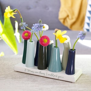 May include: A white ceramic vase with the text "If Mugs were flowers I would pick you x" and seven small colourful bud vases holding various spring flowers, including yellow daffodils, pink daisies, and blue muscari.