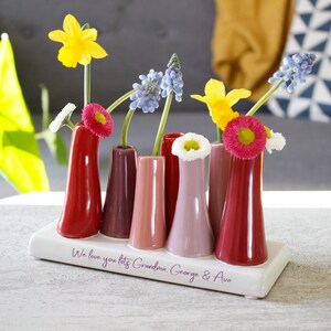 May include: A white ceramic vase with six small red, pink and purple bud vases. The vase has the inscription "We love you lots Grandma, George & Ava".
