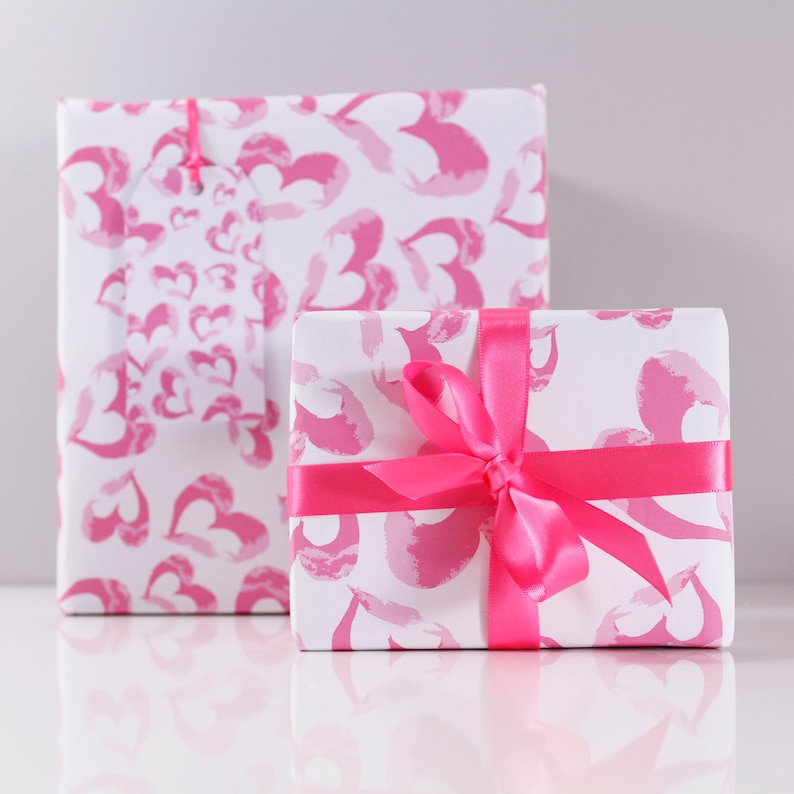 May include: Two gift boxes wrapped in white paper with a pink heart pattern. The smaller box has a pink satin ribbon tied in a bow.