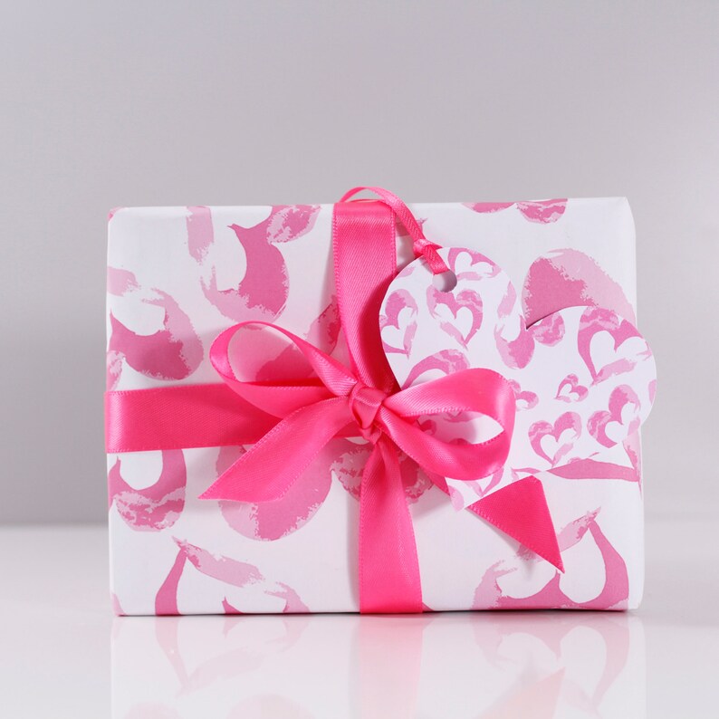 May include: A white gift box with a pink and white pattern of hearts and swirls. The box is tied with a pink satin ribbon and has a pink heart-shaped gift tag.
