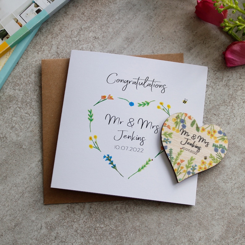 May include: A white card with a floral wreath and the text "Congratulations Mr & Mrs Jenkins 10.07.2022". A wooden heart with a floral design and the text "Mr & Mrs Jenkins 10.07.2022" is attached to the card.