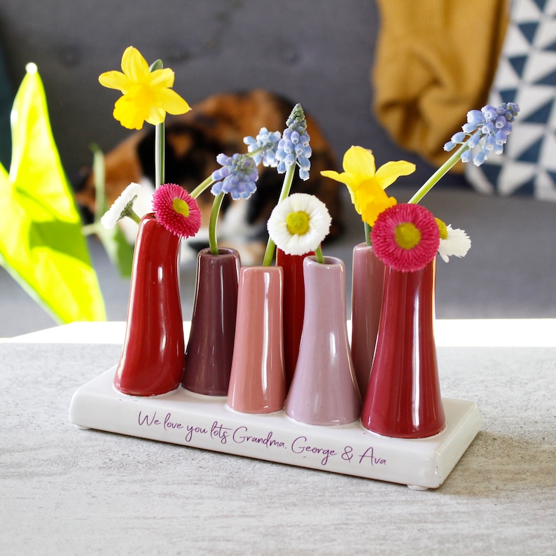 May include: A white ceramic vase with six small red, pink and purple bud vases. The vase has a message that reads "We love you lots Grandma George & Ava".