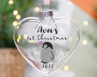 Op de afbeelding: Helder glazen kerstversiering in de vorm van een hart met een pinguïn en de tekst "Ava's 1st Christmas 2022".