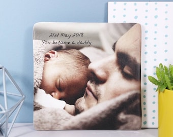 May include: Square photo print featuring a sleeping baby nestled against a person's face. The text "21st May 2018 You became a daddy" is handwritten on the print. The background includes a light blue wall and a decorative element.
