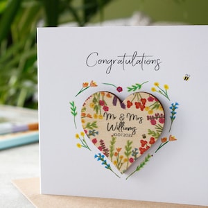 May include: A white card with the word "Congratulations" written in black cursive. A wooden heart with a floral pattern and the text "Mr. & Mrs. Williams 10.07.2022" is attached to the card.