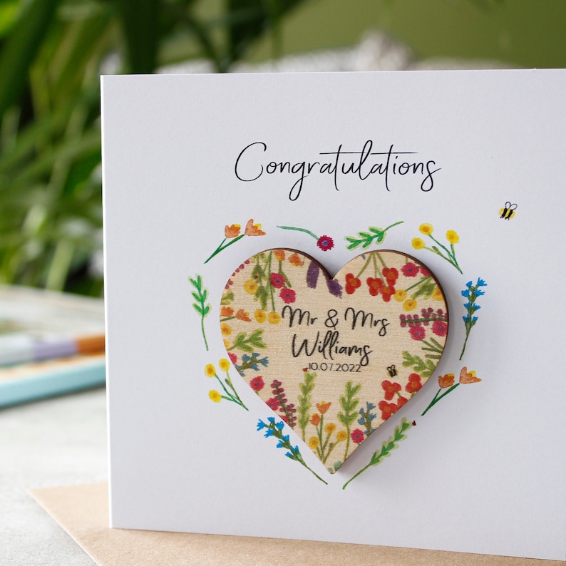 May include: A white card with the word "Congratulations" written in black cursive. A wooden heart with a floral pattern and the text "Mr. & Mrs. Williams 10.07.2022" is attached to the card.