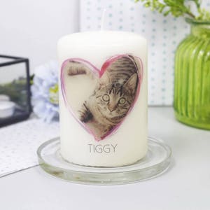 Pu&ograve; includere: Una candela a colonna bianca con un'immagine a forma di cuore di un gatto tigrato. Il gatto &egrave; all'interno di un contorno a forma di cuore rosa. Il nome "TIGGY" &egrave; stampato sotto. La candela poggia su un piatto di vetro trasparente.