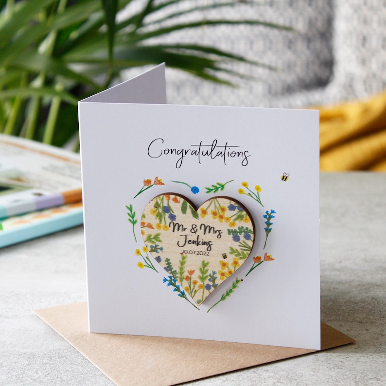 May include: A white card with the word "Congratulations" printed on it. A wooden heart with a floral design and the text "Mr & Mrs Jenkins 10.07.2022" is attached to the card.