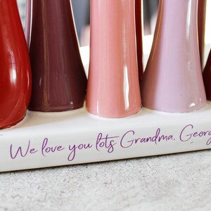 May include: A white ceramic vase with a row of colourful ceramic flowers. The vase has the text "We love you lots Grandma. Georg" written on the bottom.